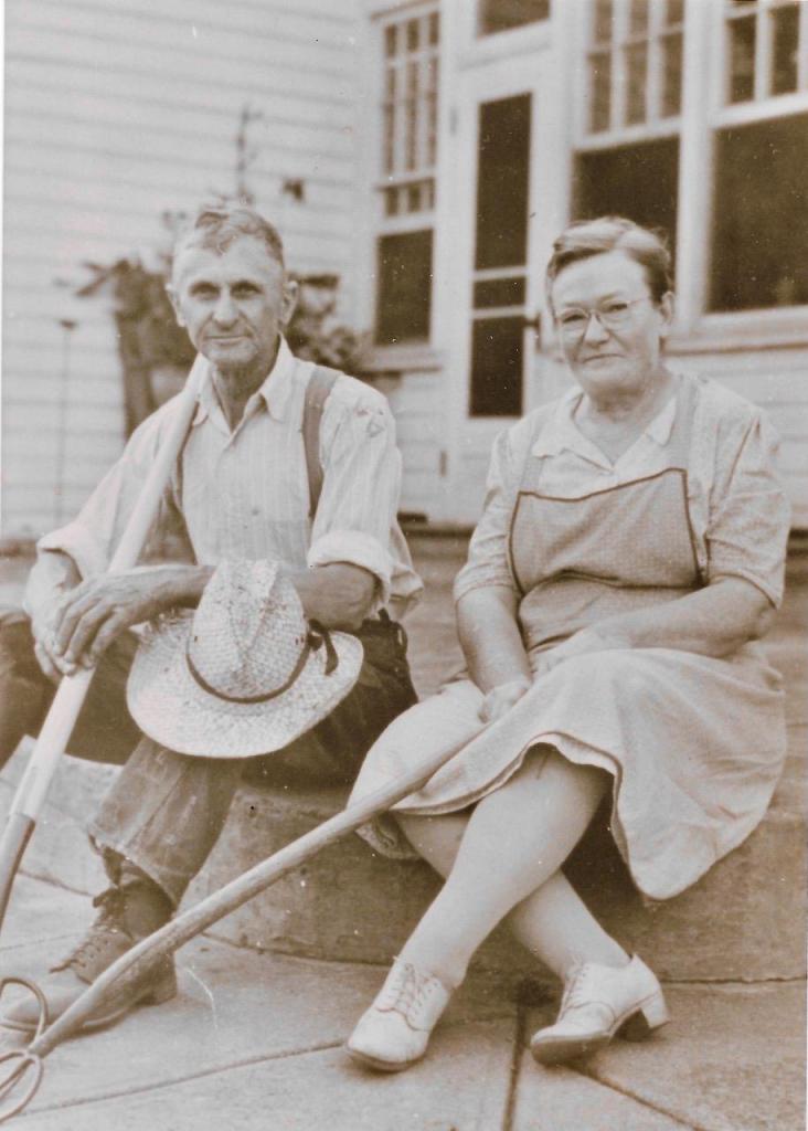 Grace and Carroll Raver, seated on a concrete step outside a house.  Carroll holds a shovel and a straw hat, Grace holds a rake.