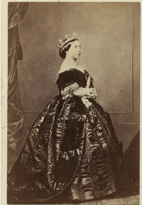 Photograph of Queen Victoria, 1861.  Queen Victoria stands facing right with her arms crossed in front of her.  She wears an elaborate dress and a crown.