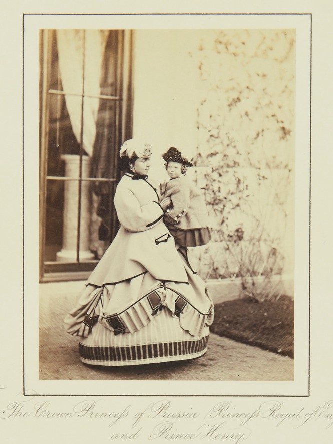 A woman wearing a dress with a hoop skirt holds a toddler and poses in front of a window.