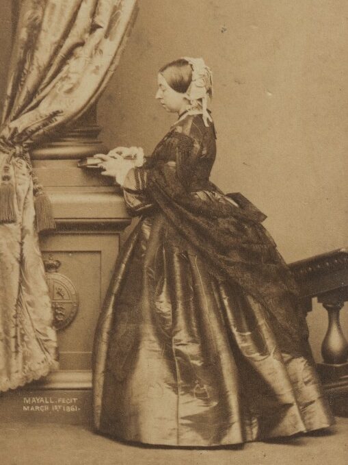 Photograph of Queen Victoria, 1861. Queen Victoria is wearing a dress and shawl, holding a book, and standing facing a column with her face in profile.