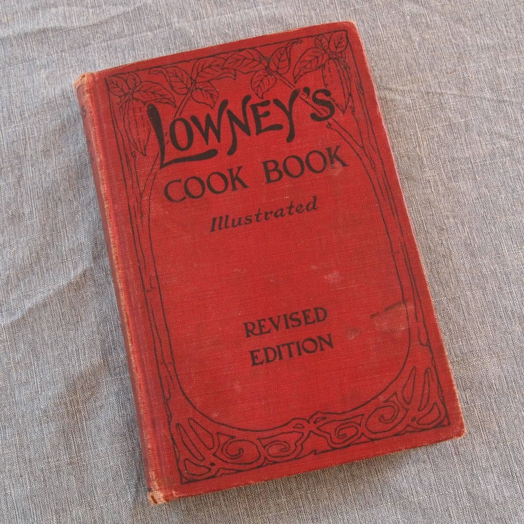 Red book with stylized illustration of cocoa trees on cover. Cover text reads "Lowney's Cook Book illustrated revised edition."